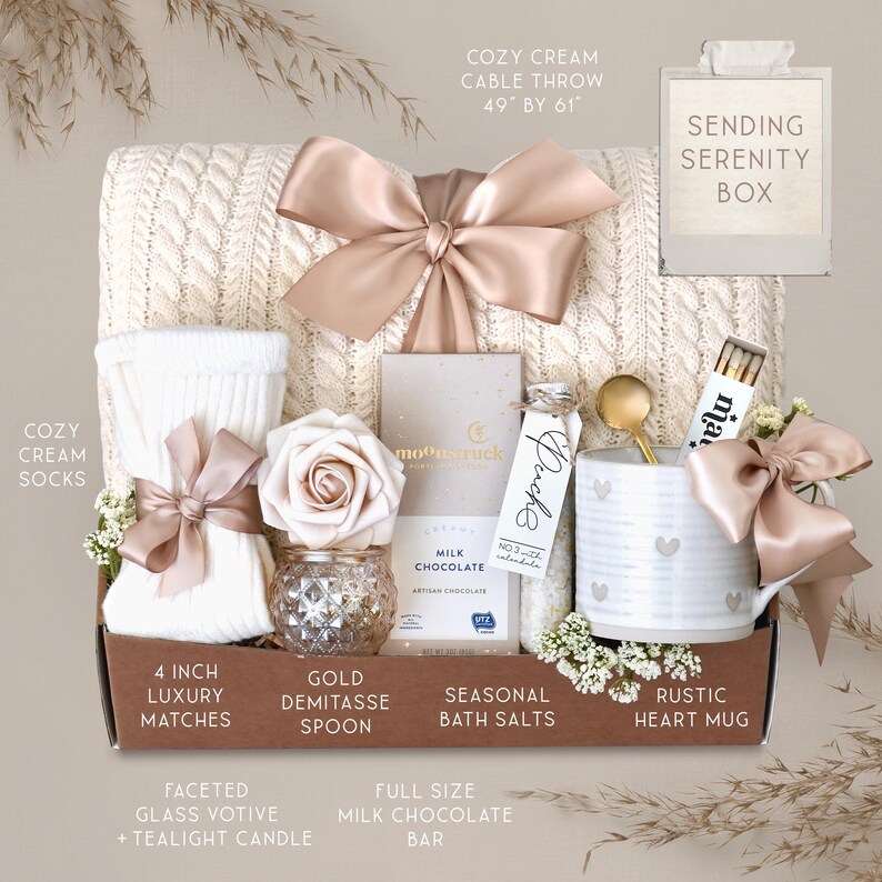 May include: A gift box with a cream-colored cable knit throw (49" x 61"), socks, a mug with hearts, a chocolate bar, bath salts, a candle, matches, and a gold spoon. The box is tied with a satin bow.