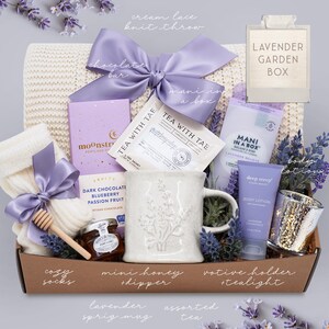 May include: A gift basket with a cream-colored knit throw, lavender-themed items, and a light purple bow. Includes a mug, body lotion, tea, chocolate, cozy socks, honey, and a votive holder. The box is labeled "Lavender Garden Box."
