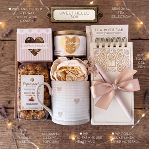 May include: A gift box labeled Sweet Hello Box containing a hand-poured soy wax candle, a bag of caramel popcorn, a ceramic mug with heart patterns, a faux peony, a tea selection, a spiral-bound notepad, and a soft-touch gel pen with a ribbon.