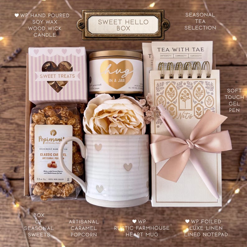 May include: A gift box labeled Sweet Hello Box containing a hand-poured soy wax candle, a bag of caramel popcorn, a ceramic mug with heart patterns, a faux peony, a tea selection, a spiral-bound notepad, and a soft-touch gel pen with a ribbon.
