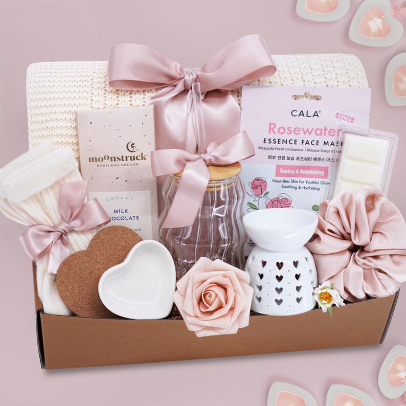 May include: A gift basket filled with self-care items. Includes a white knit scarf, a jar with a pink bow, a face mask, a white oil diffuser, a scrunchie, a heart-shaped cork coaster, a heart-shaped dish, a rose, and a chocolate bar.