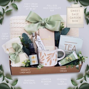 May include: A gift box with a cream cable throw (49" x 61"), socks, a mug with leaf design, tea sampler, hand cream, bath salts, and a milk chocolate bar. The box includes a Serenity Linen Mist and is decorated with a sage green bow.