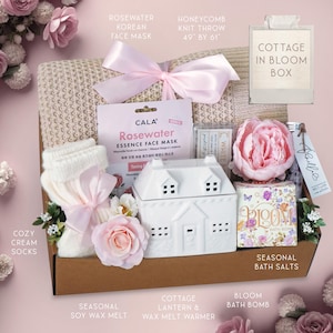 May include: A gift box filled with self-care items. Includes a honeycomb knit throw (49" x 61"), face mask, cozy cream socks, a cottage lantern, seasonal soy wax melt, bath salts, and a bloom bath bomb. Decorated with pink flowers and ribbons.