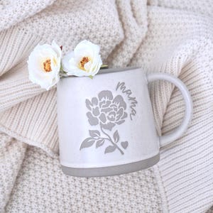 May include: A speckled light gray ceramic mug featuring a dark gray floral graphic and the word "mama" in cursive script. Two white roses with yellow centers are tucked into the mug, which has a smooth handle and a solid gray base.