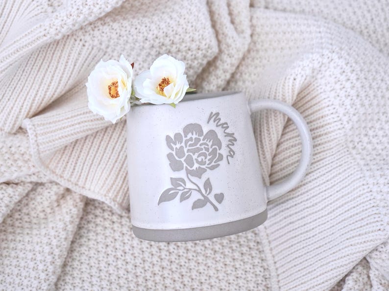 May include: A speckled light gray ceramic mug featuring a dark gray floral graphic and the word "mama" in cursive script. Two white roses with yellow centers are tucked into the mug, which has a smooth handle and a solid gray base.
