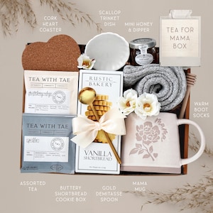 May include: A gift box containing a heart-shaped cork coaster, a white scallop trinket dish, a jar of honey with a dipper, a Tea for Mama box, two Tea with Tae sachets, a box of vanilla shortbread, a gold spoon, gray socks, and a floral mug.