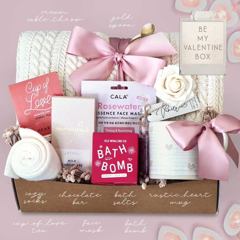 May include: A Valentine's Day gift box with a cream cable knit throw, pink bow, and various self-care items. Includes a mug, bath bomb, face mask, chocolate bar, tea, and cozy socks. The box is labeled "Be My Valentine Box."