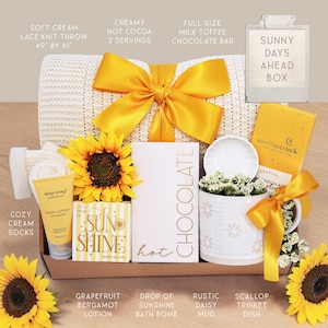 May include: A gift box containing a cream lace knit throw with a yellow ribbon, a Moonstruck milk toffee chocolate bar, a white mug with daisy patterns, a small white dish, a Deep Steep grapefruit bergamot lotion, and a Sun Shine bath bomb.