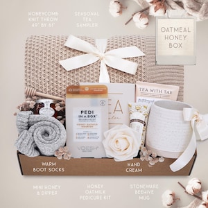 May include: A gift box with a beige honeycomb knit throw (49" x 61"), a white bow, and various self-care items. Includes a mug, socks, honey, a pedicure kit, and hand cream. The box is tied with a white ribbon.