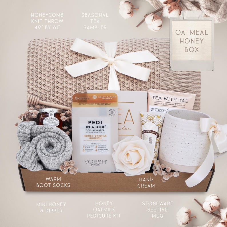 May include: A gift box with a beige honeycomb knit throw (49" x 61"), a white bow, and various self-care items. Includes a mug, socks, honey, a pedicure kit, and hand cream. The box is tied with a white ribbon.