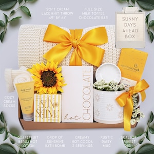 May include: A gift box with a soft cream lace knit throw (49" x 61"), a sunflower, and a yellow bow. Includes a chocolate bar, lotion, bath bomb, hot cocoa, a daisy mug, and a trinket dish. The box is labeled "Sunny Days Ahead".