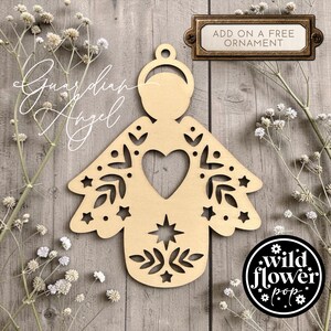 May include: A light-colored wooden angel ornament featuring intricate laser-cut details including a central heart, stars, and leaf patterns. The ornament has a circular loop at the top for hanging. Text on the image reads Guardian Angel and Add on a free ornament.