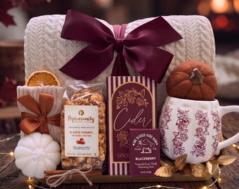 May include: A gift set featuring a cream-colored cable knit blanket with a large burgundy bow, a white pumpkin-shaped mug with floral patterns, a bag of Popinsanity caramel popcorn, a box of cider, a blackberry goat milk soap, and a white pumpkin candle.