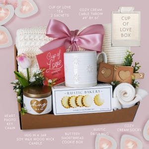 May include: A gift basket with a cream-colored cable knit throw, a white daisy-patterned mug, a box of vanilla shortbread cookies, and a candle labeled "hug in a jar." Includes a "Cup of Love" tea and a small photo holder.