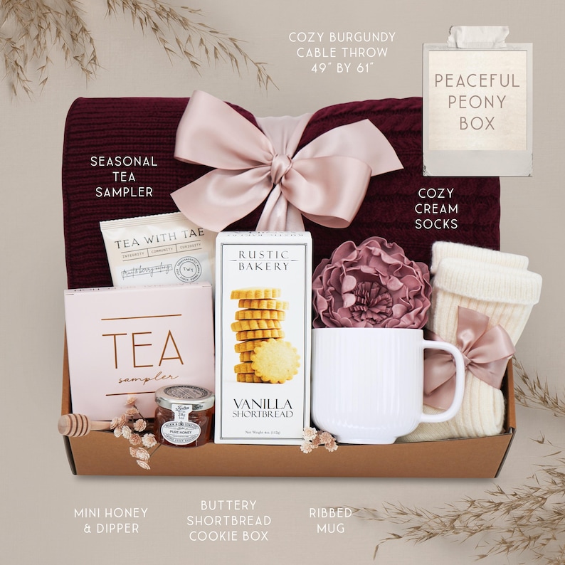 May include: A gift box filled with cozy items. Includes a burgundy cable throw (49" by 61"), cream socks, a tea sampler, vanilla shortbread cookies, a ribbed mug, and mini honey with a dipper. A pink bow adorns the box.