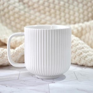 May include: A white ceramic mug featuring a vertical ribbed texture on the exterior surface. The mug has a smooth, rounded handle on the left side and a clean, cylindrical shape. The finish is matte white, providing a consistent and uniform appearance.