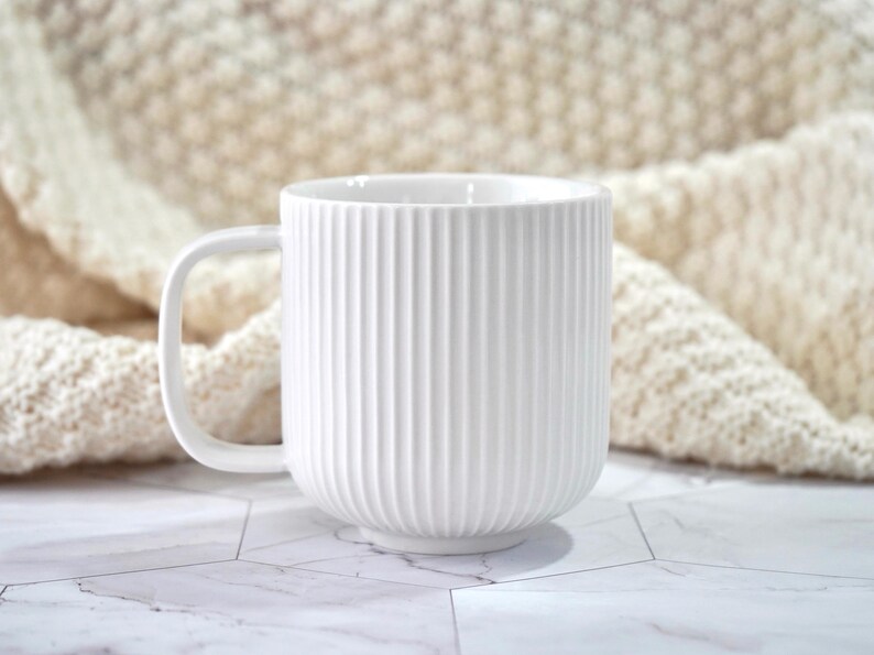 May include: A white ceramic mug featuring a vertical ribbed texture on the exterior surface. The mug has a smooth, rounded handle on the left side and a clean, cylindrical shape. The finish is matte white, providing a consistent and uniform appearance.