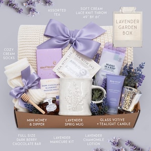 K&ouml;nnte beinhalten: Eine Geschenkbox mit Lavendel-Artikeln. Sie enth&auml;lt eine cremefarbene Strickdecke (124 cm x 155 cm), eine Tasse mit Zweig-Motiv, Tee, Lotion, eine Kerze, Socken, eine dunkle Schokoladenriegel und ein Manik&uuml;re-Set. Die Box ist mit einer lila Schleife gebunden.