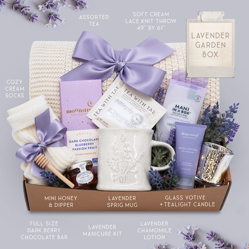 K&ouml;nnte beinhalten: Eine Geschenkbox mit Lavendel-Artikeln. Sie enth&auml;lt eine cremefarbene Strickdecke (124 cm x 155 cm), eine Tasse mit Zweig-Motiv, Tee, Lotion, eine Kerze, Socken, eine dunkle Schokoladenriegel und ein Manik&uuml;re-Set. Die Box ist mit einer lila Schleife gebunden.