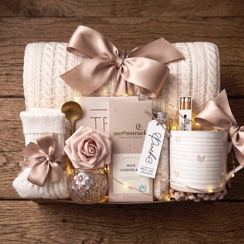 Op de afbeelding: Een geschenkmand met een cr&egrave;mekleurige kabelgebreide deken met een grote satijnen strik, witte geribbelde sokken met een strik, een gouden lepel, een kunstroos in een glazen pot, een Moonstruck melkchocoladereep, een flesje met label en een hartjesmok.