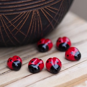 Könnte beinhalten: Sechs rote und schwarze Marienkäfer-Perlen auf einer Holzoberfläche. Die Perlen sind glänzend und haben ein realistisches Marienkäfer-Design.