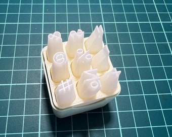 Könnte beinhalten: Ein weißer Plastikbehälter mit neun verschiedenen weißen Plastik-Spritzdüsen. Die Düsen haben verschiedene Designs, darunter einen Stern, einen Wirbel, einen Kreis, ein Nummernschild, ein Prozentzeichen und ein Pluszeichen.