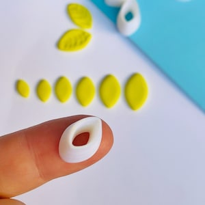 May include: A collection of white and yellow polymer clay leaf-shaped cutters and leaves. The cutters are in various sizes and shapes, with a white oval cutter held by a finger. The leaves are yellow and arranged in a row.