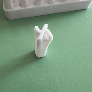 May include: A white, 3D-printed object with a unique, abstract shape, possibly a tool or component. The object is positioned on a light green surface, with a matching white tray in the background containing similar items. The design features a central, upward-pointing form with multiple extensions.