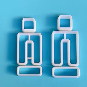 May include: Two white plastic cookie cutters in the shape of a person, with a rectangular head, torso, and feet. The cutters are on a solid light blue background. The cookie cutters are for baking.
