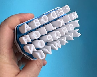Könnte beinhalten: Ein blaues und weißes Alphabet-Stempelset aus Kunststoff mit einzelnen Stempeln für jeden Buchstaben des Alphabets. Die Stempel sind in einem Raster angeordnet und werden in einer Hand gehalten.