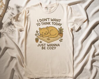 Peut inclure: Un t-shirt crème avec un motif représentant un chat dormant sur un oreiller entouré de plantes. Le texte sur le t-shirt est "I don't want to think today just wanna be cozy".