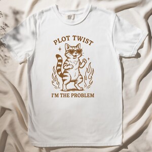 Puede incluir: Camiseta blanca con una ilustración marrón de un gato con gafas de sol de pie en llamas. El texto "Plot Twist I'm the Problem" está impreso debajo del gato.