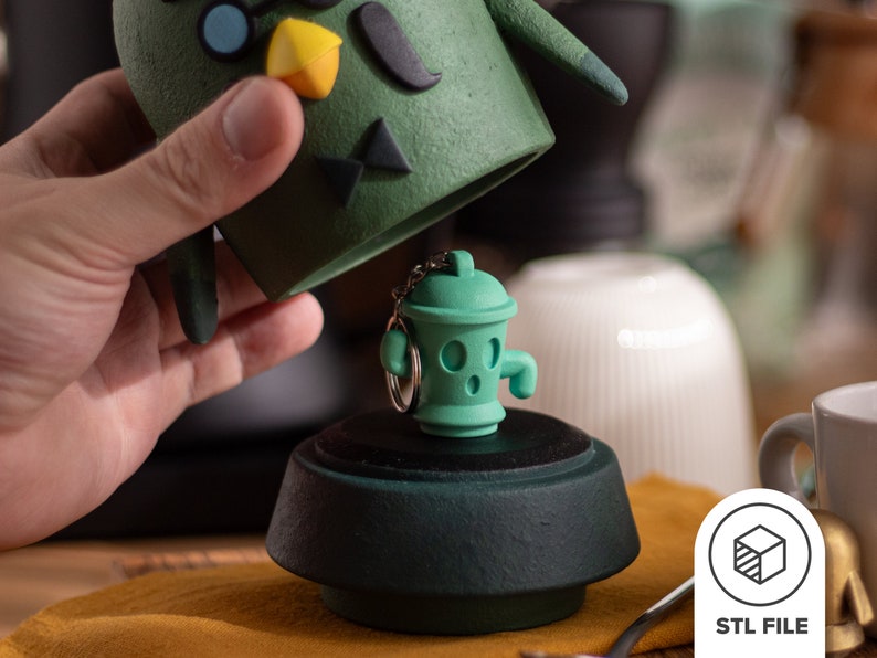 Pu&ograve; includere: Una statuetta di ceramica verde di un personaggio dei cartoni animati con un portachiavi attaccato. La statuetta ha la forma di un idrante con un viso e delle braccia. Il portachiavi &egrave; attaccato alla parte superiore della testa della statuetta. La statuetta &egrave; seduta su una base verde.