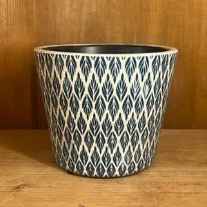 May include: A ceramic plant pot with a navy blue and white leaf pattern. The pot has a tapered shape and a dark interior. The pot is sitting on a wooden surface.
