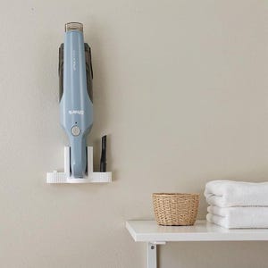 Puede incluir: Una aspiradora de mano Shark azul claro montada en un soporte de pared blanco. La aspiradora está en posición vertical, con un pequeño accesorio de boquilla. Una pequeña cesta tejida y toallas blancas dobladas están en un estante blanco debajo.