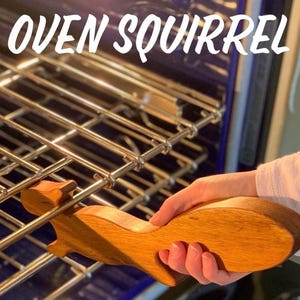 May include: A wooden squirrel-shaped oven rack puller is held in front of an oven rack. The text "OVEN SQUIRREL" is displayed in white, cursive font above the rack puller. The puller is a light brown colour.