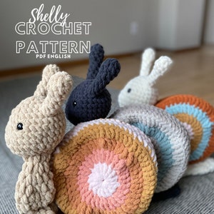 May include: A crochet pattern for making a snail with bunny ears. The snail is made in a rainbow of colours, including orange, pink, white, blue, and grey. The bunny ears are beige and black.