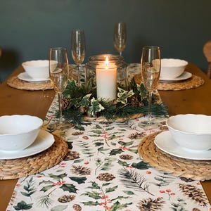 May include: A wooden table set for a festive dinner with a white tablecloth featuring a green and red floral pattern. The table is set with white bowls, plates, and glasses. A candle is in the centre of the table surrounded by greenery.