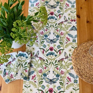 May include: A white table runner with a green, blue and pink floral pattern featuring butterflies. The runner is on a wooden table with a woven placemat and a bouquet of green plants.
