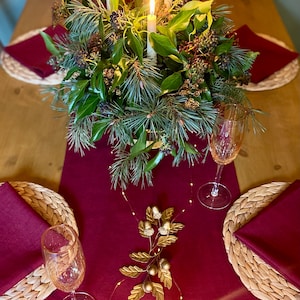May include: A Christmas table setting with a burgundy runner, napkins, and woven placemats. A centerpiece of greenery and lit candles sits on the table. Two champagne glasses and a gold leaf and acorn decoration complete the festive scene.