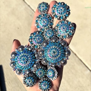 Könnte beinhalten: Eine Sammlung kreisförmiger, silberfarbener Ornamente im Concho-Stil mit gewelltem Rand und einem zentralen Cluster aus irisierenden blauen und klaren Kristallen. Jedes Stück hat ein metallisches Finish mit kleinen, erhabenen silbernen Nieten.