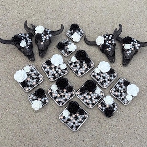May include: A collection of two black bull skull wall hangings and twelve square decorative tiles, all embellished with a mix of black and white floral accents, small black beads, and iridescent white pearls arranged in a dense, textured pattern.