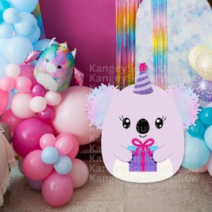 May include: A purple koala wearing a party hat and holding a purple gift box. The koala is surrounded by colorful balloons and a rainbow backdrop.