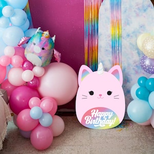 May include: A pink cardboard cutout shaped like a cat with a unicorn horn and rainbow design. The cutout reads "Happy Birthday!" in a white font. The cutout is surrounded by pink, blue and white balloons.