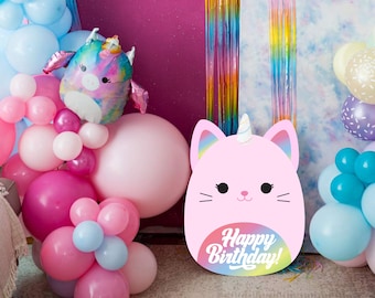 May include: A pink cat-shaped cardboard cutout with a unicorn horn and rainbow design. The cutout says "Happy Birthday!" in a white font. The cutout is surrounded by pink, blue, and white balloons.