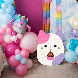 Puede incluir: Un peluche grande con forma de gato blanco que sostiene una magdalena con una vela encendida. El gato tiene detalles en rosa y morado. El fondo incluye globos rosas, azules y blancos, y un globo con forma de unicornio.