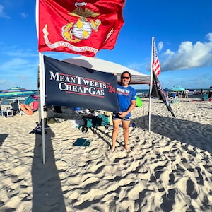May include: A person holding a black sign with the text "Mean Tweets Cheap Gas 24" on a beach. There are two flags in the background, one is a red, white, and blue American flag and the other is a red flag with the words "United States Marines" and a globe with a globe with a golden eagle on top.