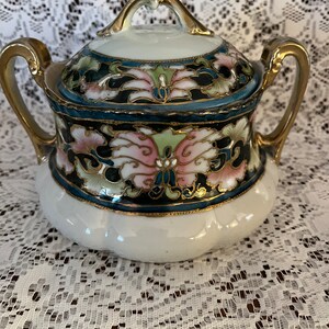 May include: A white porcelain sugar bowl with a lid, featuring a floral design in pink, green, and gold on a black background. The bowl has gold accents on the lid and handles.