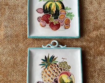 May include: Two ceramic wall plaques with a green border and a white background. The top plaque features a painted still life of a watermelon, strawberries, and grapes. The bottom plaque features a painted still life of a pineapple, grapes, and an apple.