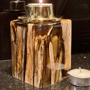 May include: A square wooden candle holder with a clear resin top and a gold metal candle cup. The holder has a natural wood grain pattern and is lit with a tea light candle.